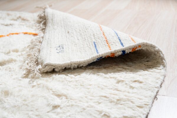 High-pile cream rug with orange accents folded over, showing a woven backing with blue and orange stripes on light wood flooring.