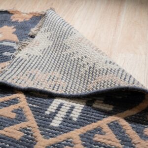 A patterned blue, tan, and white rug is lifted to show its woven backing on a light wood floor.