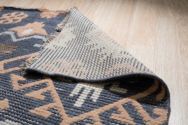 A patterned blue, tan, and white rug is lifted to show its woven backing on a light wood floor.