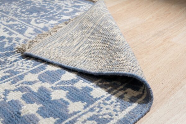 Blue geometric rug folded over, displaying the beige woven reverse side on light wood flooring.