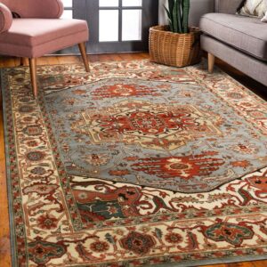 A traditional red, cream, and blue patterned rug rests on hardwood floors between a pink armchair and a gray sofa.
