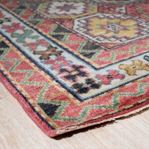 Low-pile rug with pink, teal, and yellow geometric designs rests on light wood grain floor.