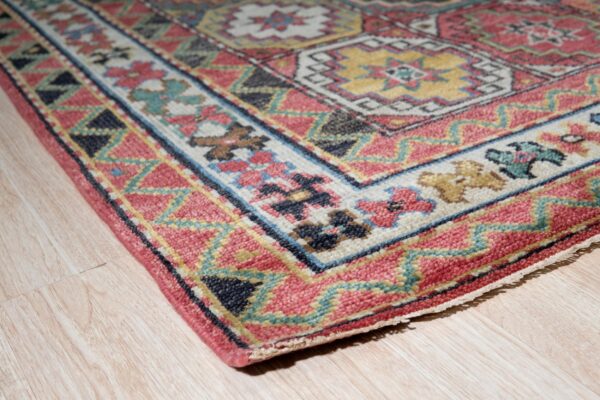 Low-pile rug with pink, teal, and yellow geometric designs rests on light wood grain floor.
