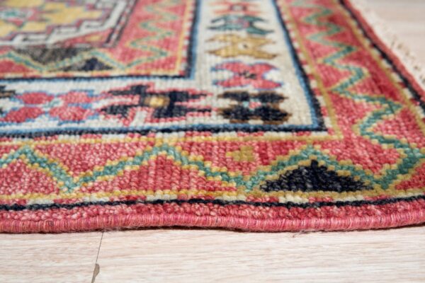 Angled view of a textured rug featuring red, blue, cream, and teal geometric borders on blonde flooring.