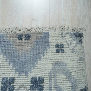 Textured cream rug with gray and blue block patterns and fringe lies on light blonde wood flooring.