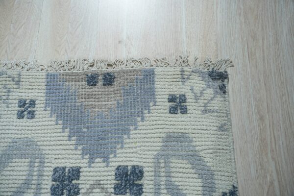 Textured cream rug with gray and blue block patterns and fringe lies on light blonde wood flooring.