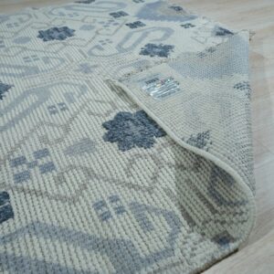 A flatweave area rug with beige, light gray, and dark blue floral patterns lies on light wood flooring.