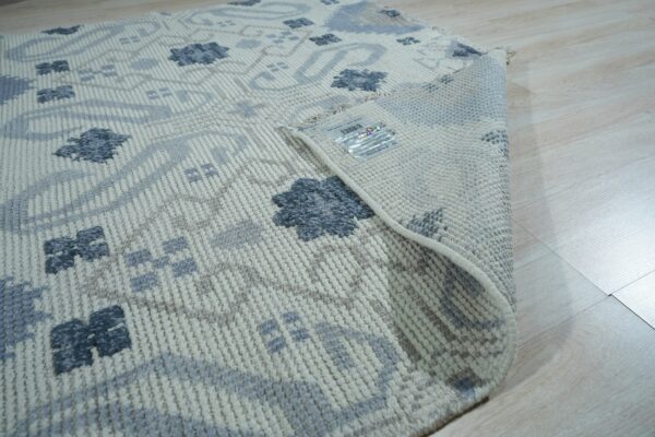 A flatweave area rug with beige, light gray, and dark blue floral patterns lies on light wood flooring.