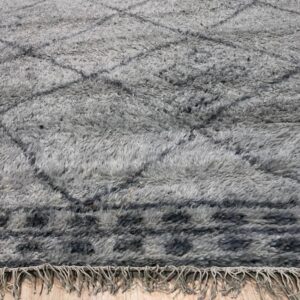 A high-pile gray rug features dark geometric patterns and short fringe lying on light wood flooring.