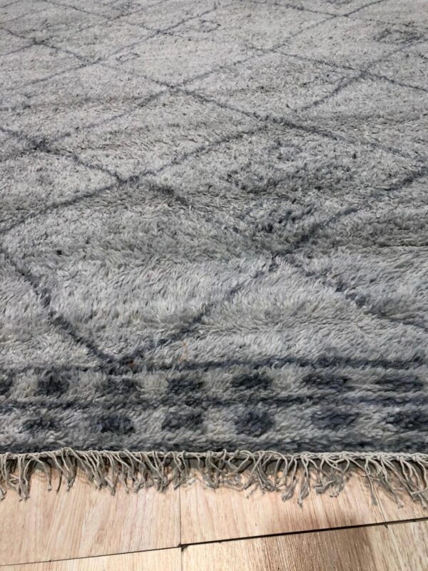 A high-pile gray rug features dark geometric patterns and short fringe lying on light wood flooring.