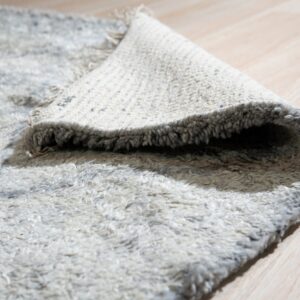 Close-up of a thick, shaggy gray rug partially rolled back, resting on pale blonde wood flooring.
