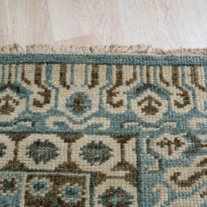 Close-up of a rug corner showing a low pile pattern in teal, brown, and beige with fringe on light wood flooring.