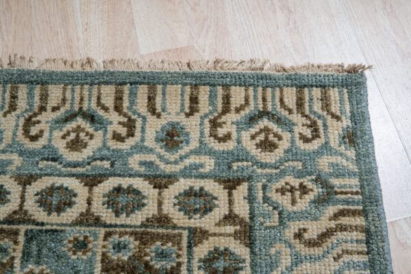 Close-up of a rug corner showing a low pile pattern in teal, brown, and beige with fringe on light wood flooring.