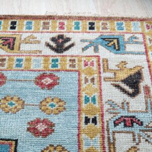 A rug corner with tribal geometric designs in blue, ivory, and gold rests on light wood planks.