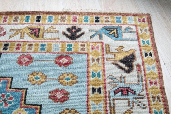 A rug corner with tribal geometric designs in blue, ivory, and gold rests on light wood planks.