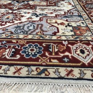 Close-up of a high-pile rug featuring intricate traditional patterns in red-brown, cream, and blue-gray on a wood floor.