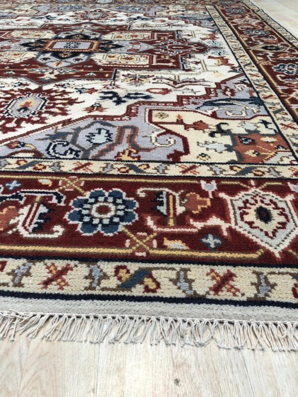 Close-up of a high-pile rug featuring intricate traditional patterns in red-brown, cream, and blue-gray on a wood floor.