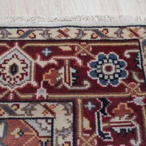 Red traditional rug corner with blue and ivory detail, fringe, and a beige border sits on light wood.