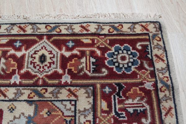 Red traditional rug corner with blue and ivory detail, fringe, and a beige border sits on light wood.
