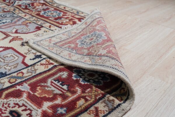 The edge of a red, blue, and cream patterned rug is rolled on light blonde wood flooring.
