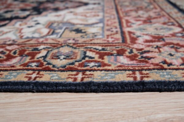 Close-up showing the navy-bound edge of a patterned rug in red, blue, and tan resting on wood floor.