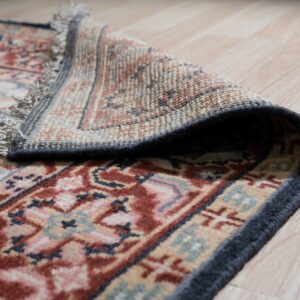 Low-pile rug with rust and navy patterns is curled back, revealing the woven backing and fringe on light wood.