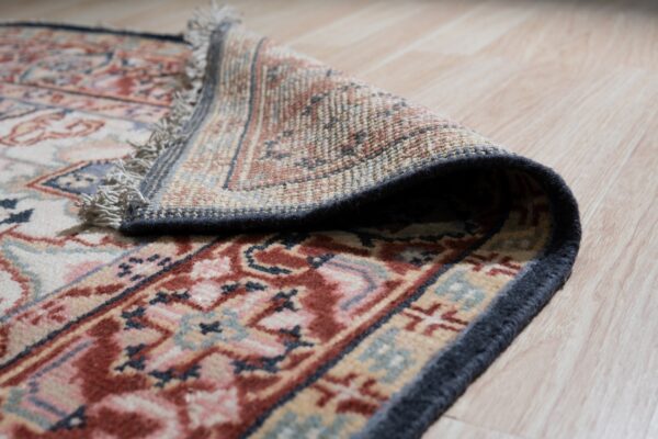 Low-pile rug with rust and navy patterns is curled back, revealing the woven backing and fringe on light wood.