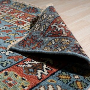 Blue, rust, and cream geometric rug detail, partially rolled up on light wood flooring.