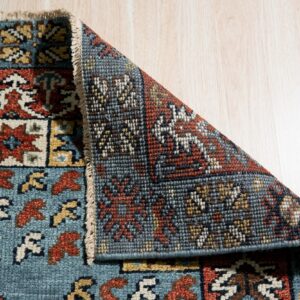 A low pile rug with teal, red, and gold geometric patterns is folded back, exposing its dense reverse side on wood flooring.