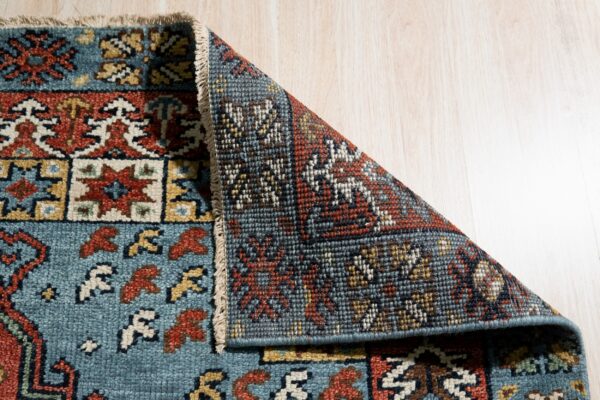 A low pile rug with teal, red, and gold geometric patterns is folded back, exposing its dense reverse side on wood flooring.