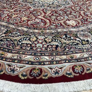 Ornate circular rug in red, navy, and cream floral patterns with white fringe on light wood floor.