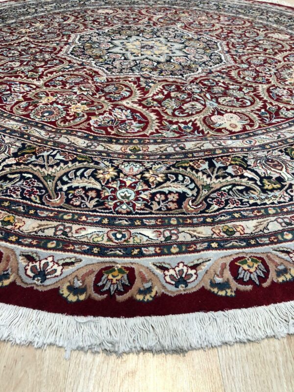 Ornate circular rug in red, navy, and cream floral patterns with white fringe on light wood floor.
