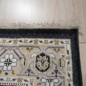 Geometric beige, black, and gray rug with medium pile and fringe detail rests on light wood flooring.