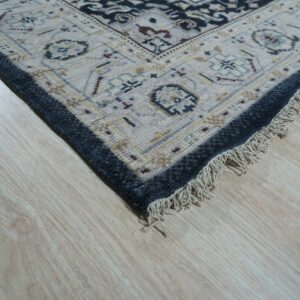 Low-pile rug in cream and charcoal with traditional patterns and fringe rests on light blonde wood floor.