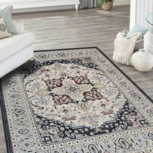 A cream and navy traditional rug with red accents lies on gray wood floors near a white sofa.