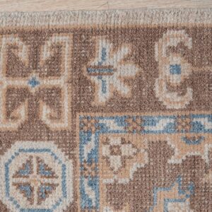 Textured pile rug in brown, beige, and light blue geometric patterns with fringe resting on a light wood floor.