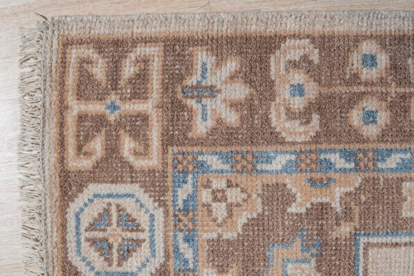 Textured pile rug in brown, beige, and light blue geometric patterns with fringe resting on a light wood floor.