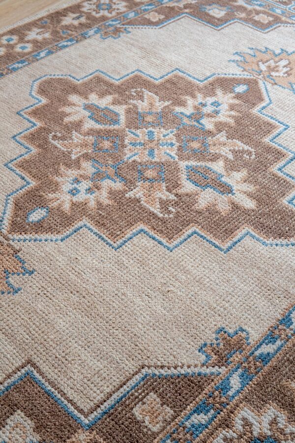 Angled view of a low-pile rug featuring geometric motifs in beige, brown, and light blue.
