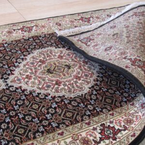 Traditional black, cream, and red medallion rug partially curled on light wood plank flooring.