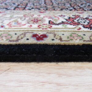 Close-up of a traditional rug border in cream, red, and black resting on light wood flooring.