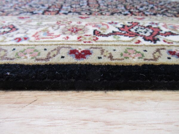 Close-up of a traditional rug border in cream, red, and black resting on light wood flooring.