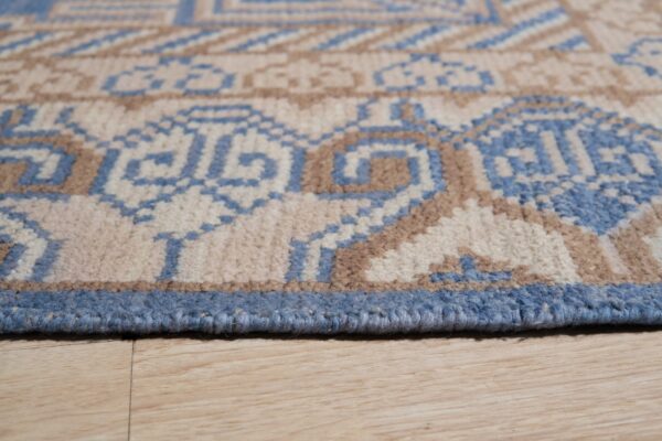 Close-up of a blue, beige, and brown geometric rug edge resting on light wood flooring.