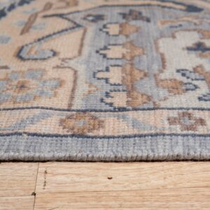 The finished edge of a low-pile rug with beige and blue geometric designs rests on light wood flooring.