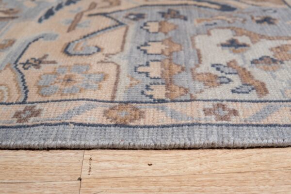 The finished edge of a low-pile rug with beige and blue geometric designs rests on light wood flooring.