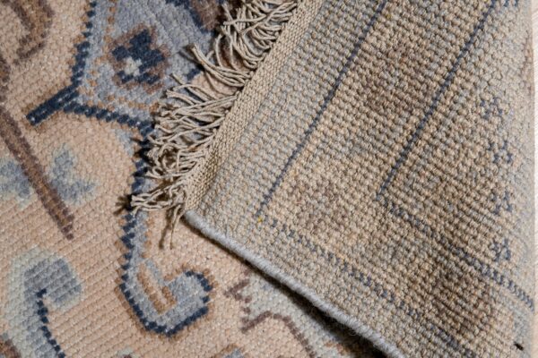 Beige rug pile with blue and brown patterns folds over showing a textured tan backing and fringe.