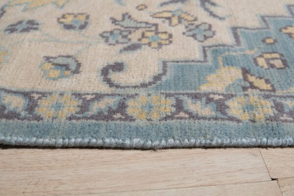 Traditional rug border detail in cream, blue, gray, and yellow resting on light wood flooring.