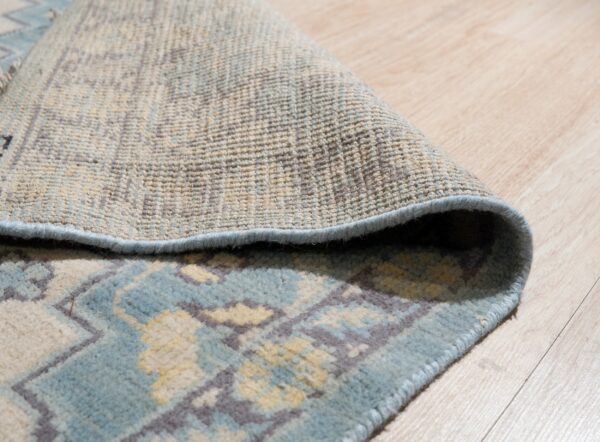 Detail of a blue and beige patterned rug curled over, showing its backing texture on light wood flooring.
