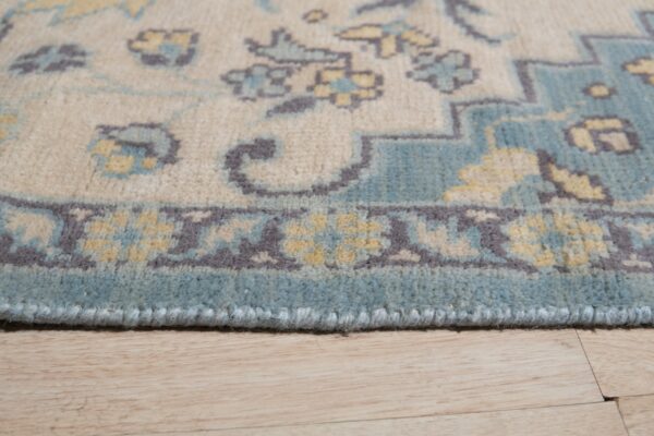The edge of a beige rug with blue, yellow, and gray traditional patterns lies on wood flooring.