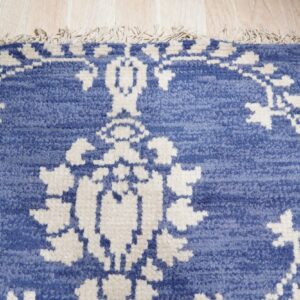 Blue textured rug with a cream traditional pattern and tassels rests on light wood planks.