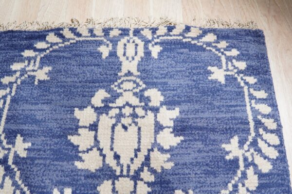 Blue textured rug with a cream traditional pattern and tassels rests on light wood planks.